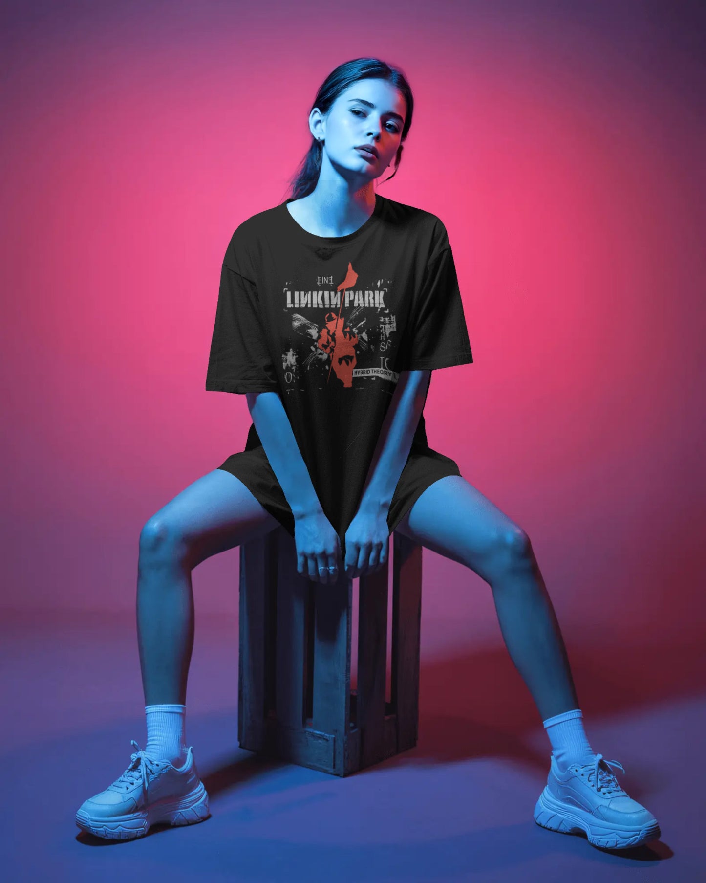 Person wearing a black t-shirt with a graphic design, sitting on a crate against a pink and blue gradient background.